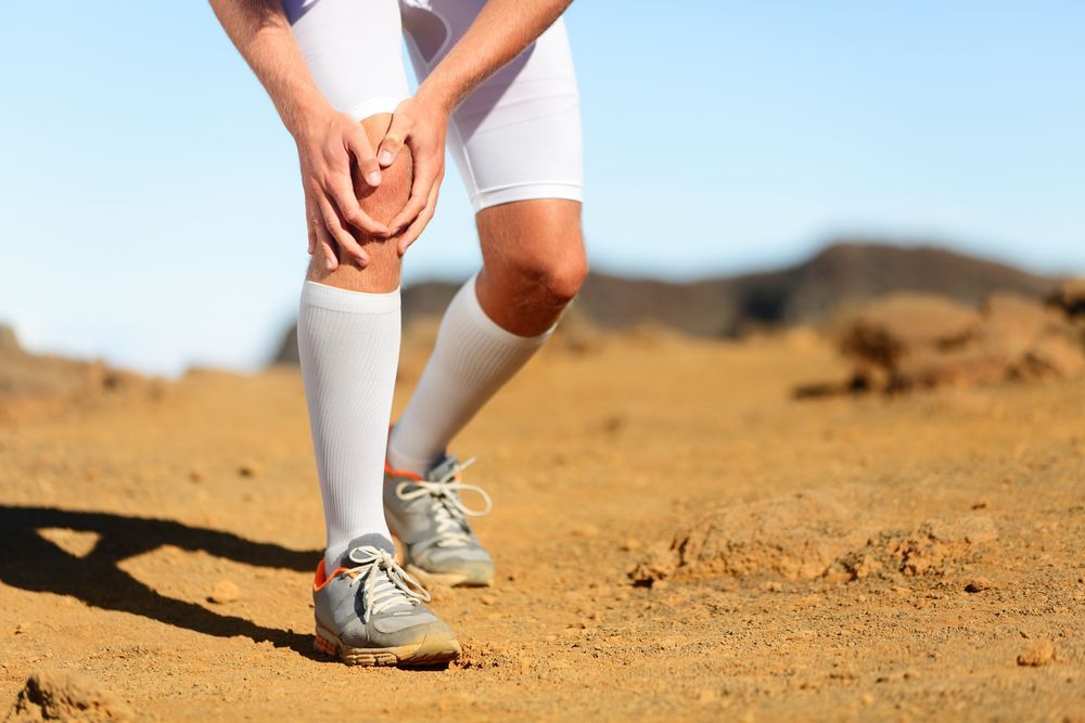 Runner holding knee in pain on a desert trail, wearing white compression gear and grey sneakers.