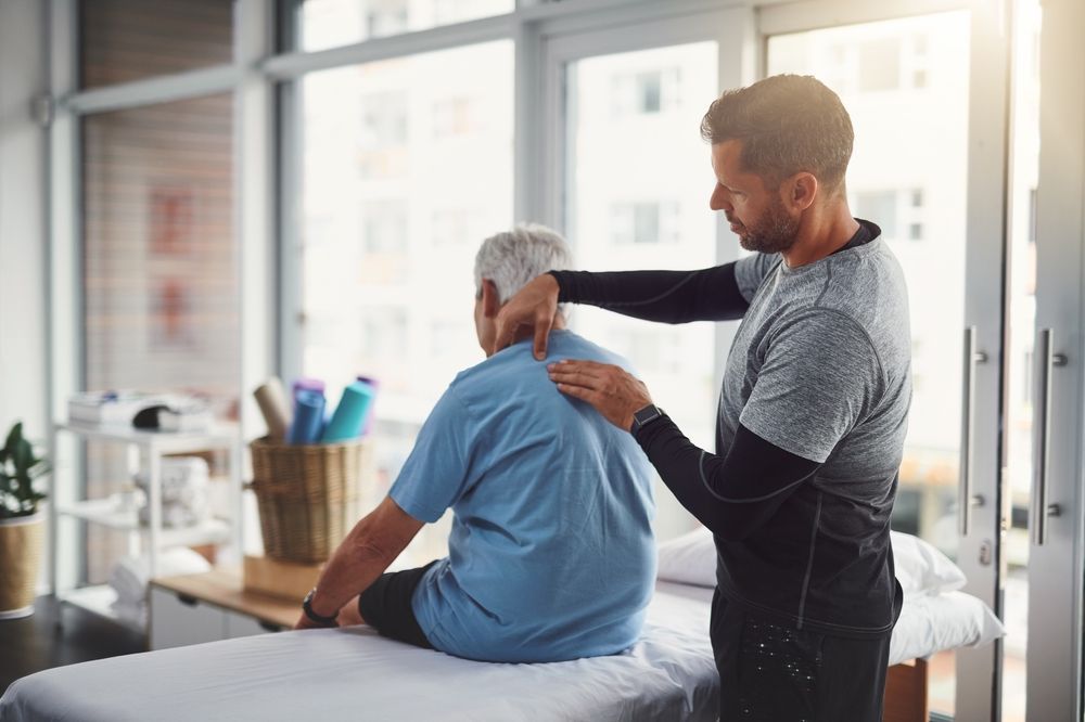 Physiotherapist examining an older patient's back in a bright office.