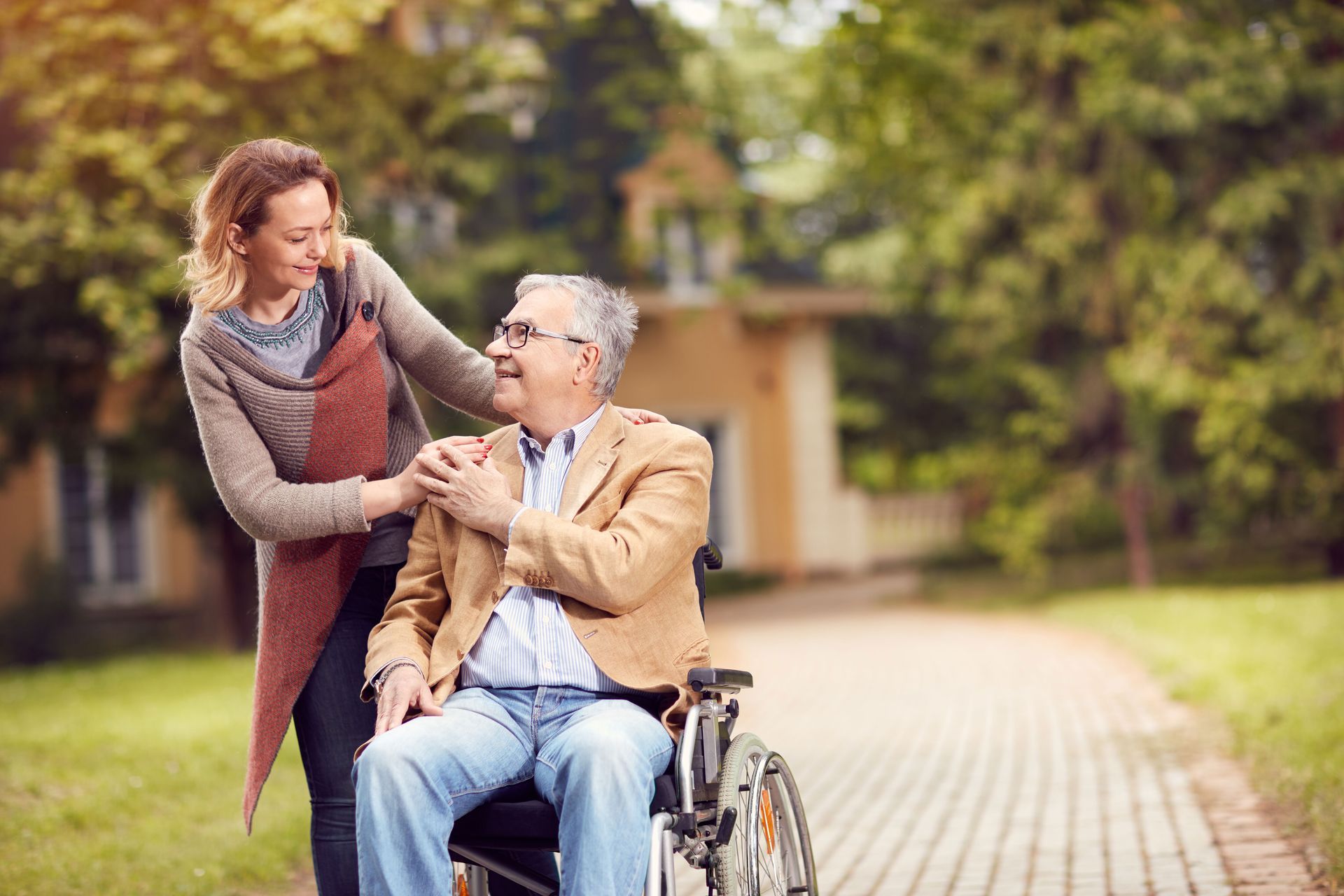 Woman helping elderly man in a wheelchair outside on a path, smiling.