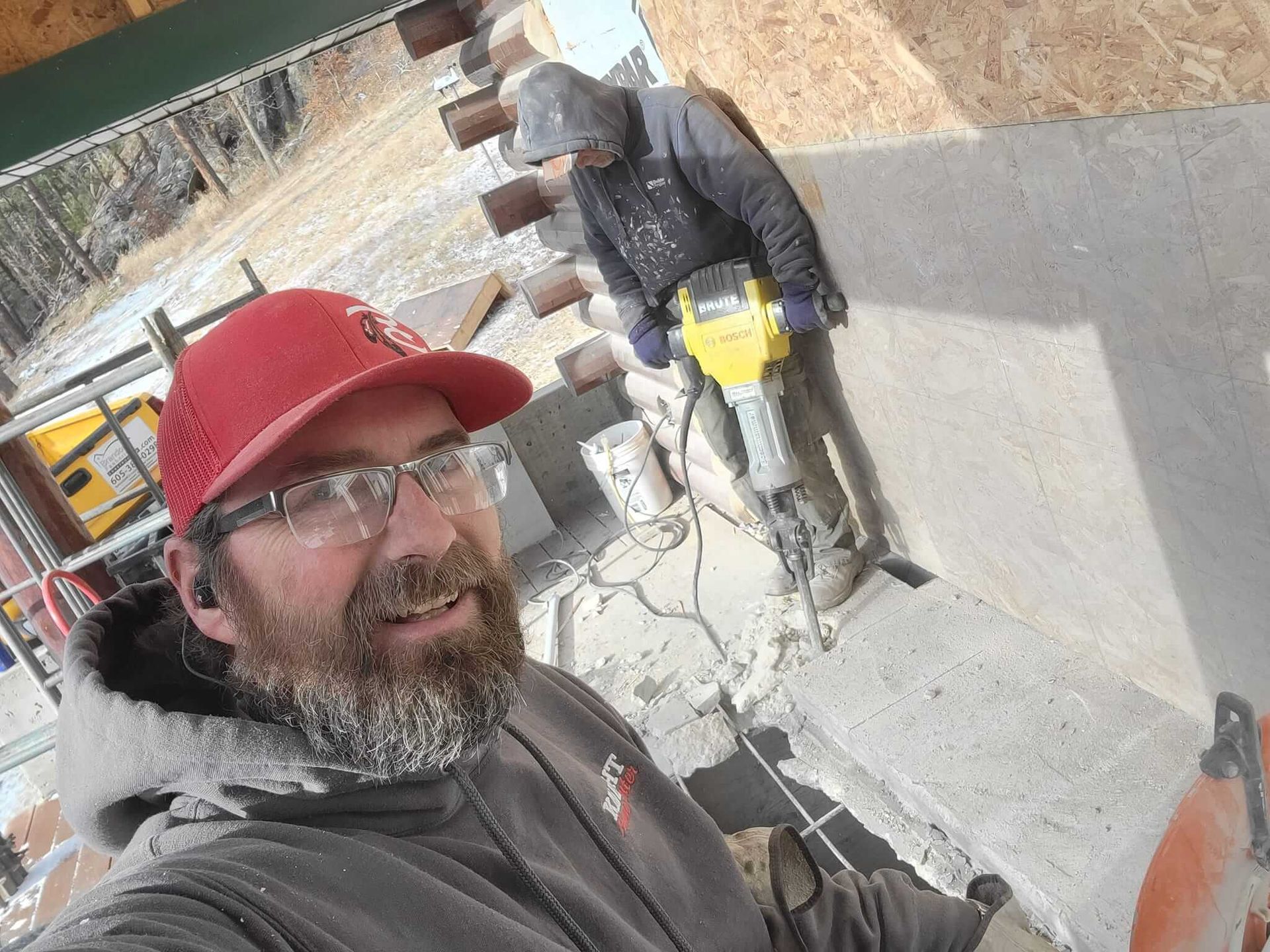 A man wearing a red hat and glasses is standing next to another man holding a hammer.