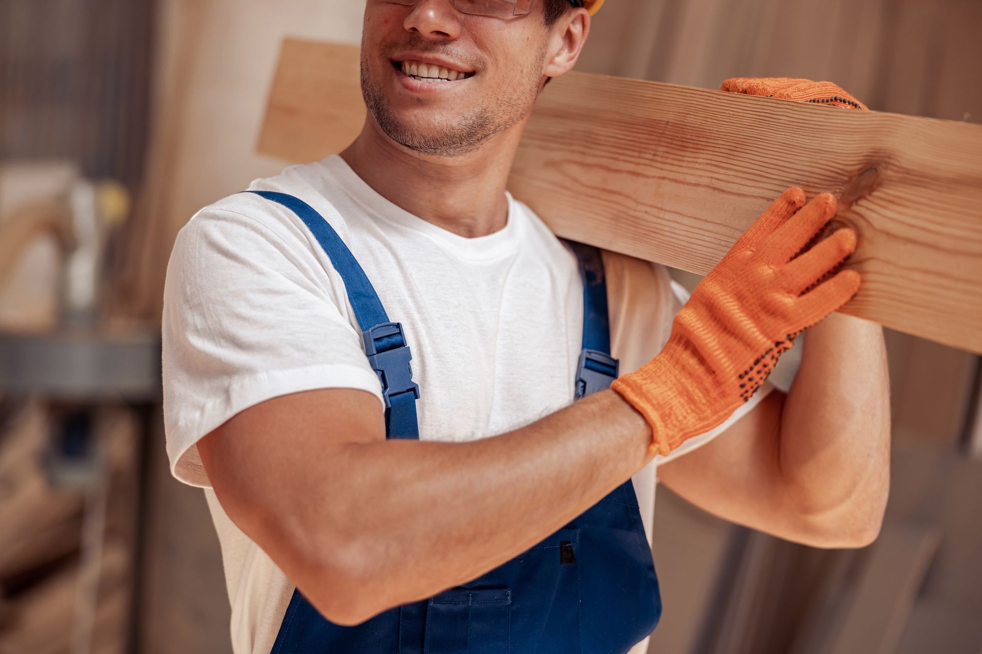 Carpenter wearing gloves and overalls, carrying wood over his shoulder, smiling.