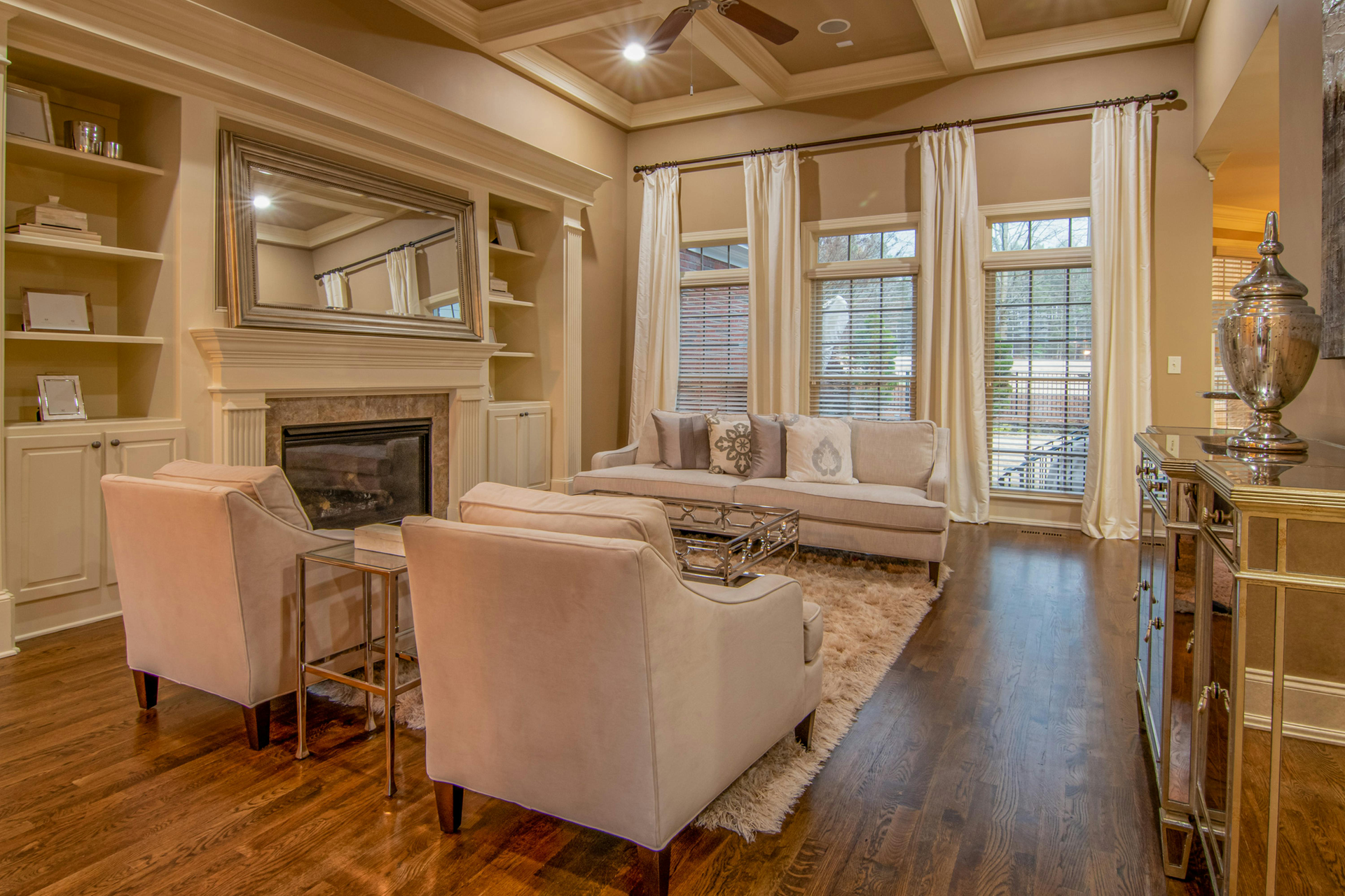 Living room with fireplace, built-in shelving, neutral-toned furniture, and large windows with cream curtains.