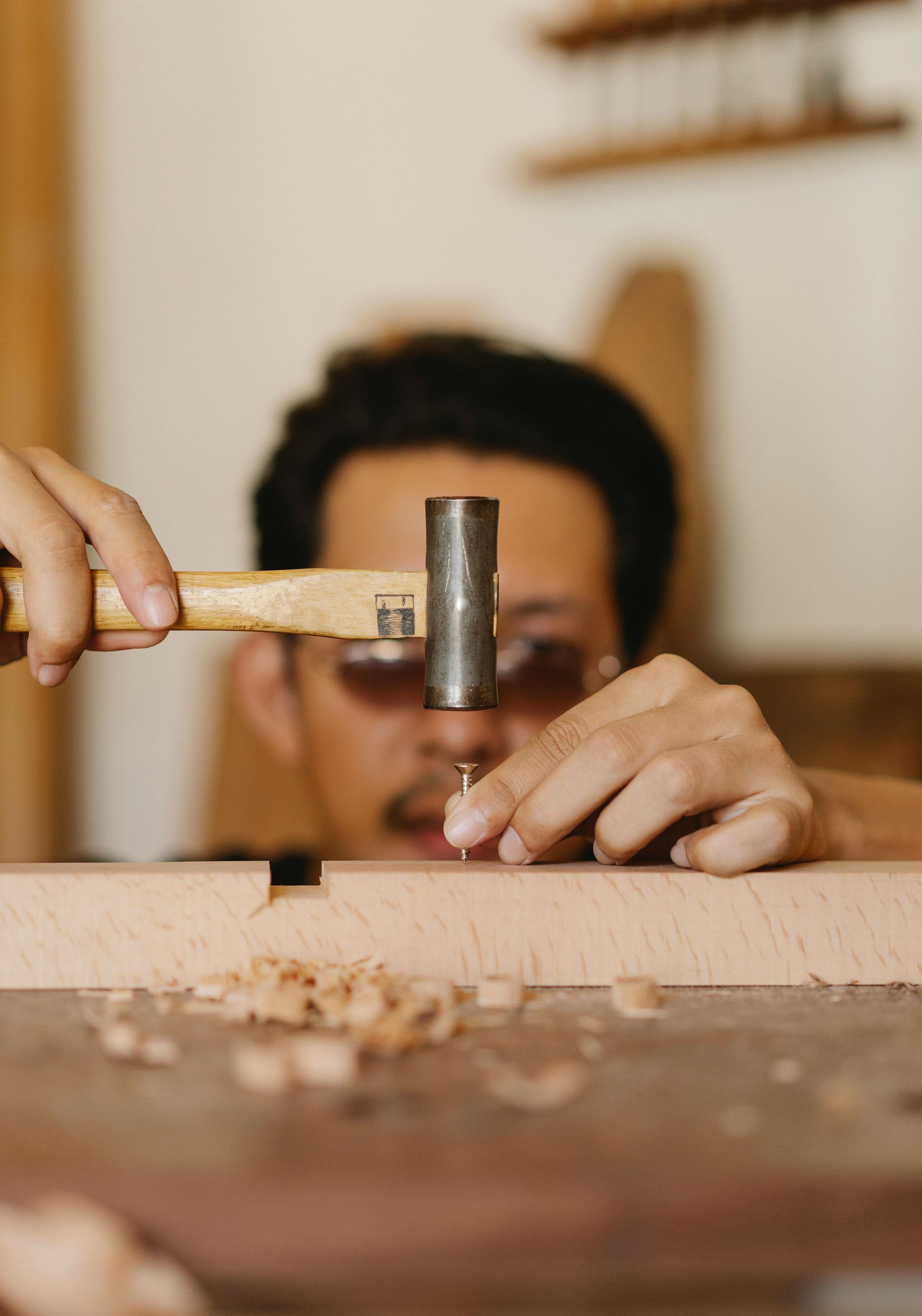A woodworker uses a hammer to drive a small nail into a piece of wood on a workshop workbench.