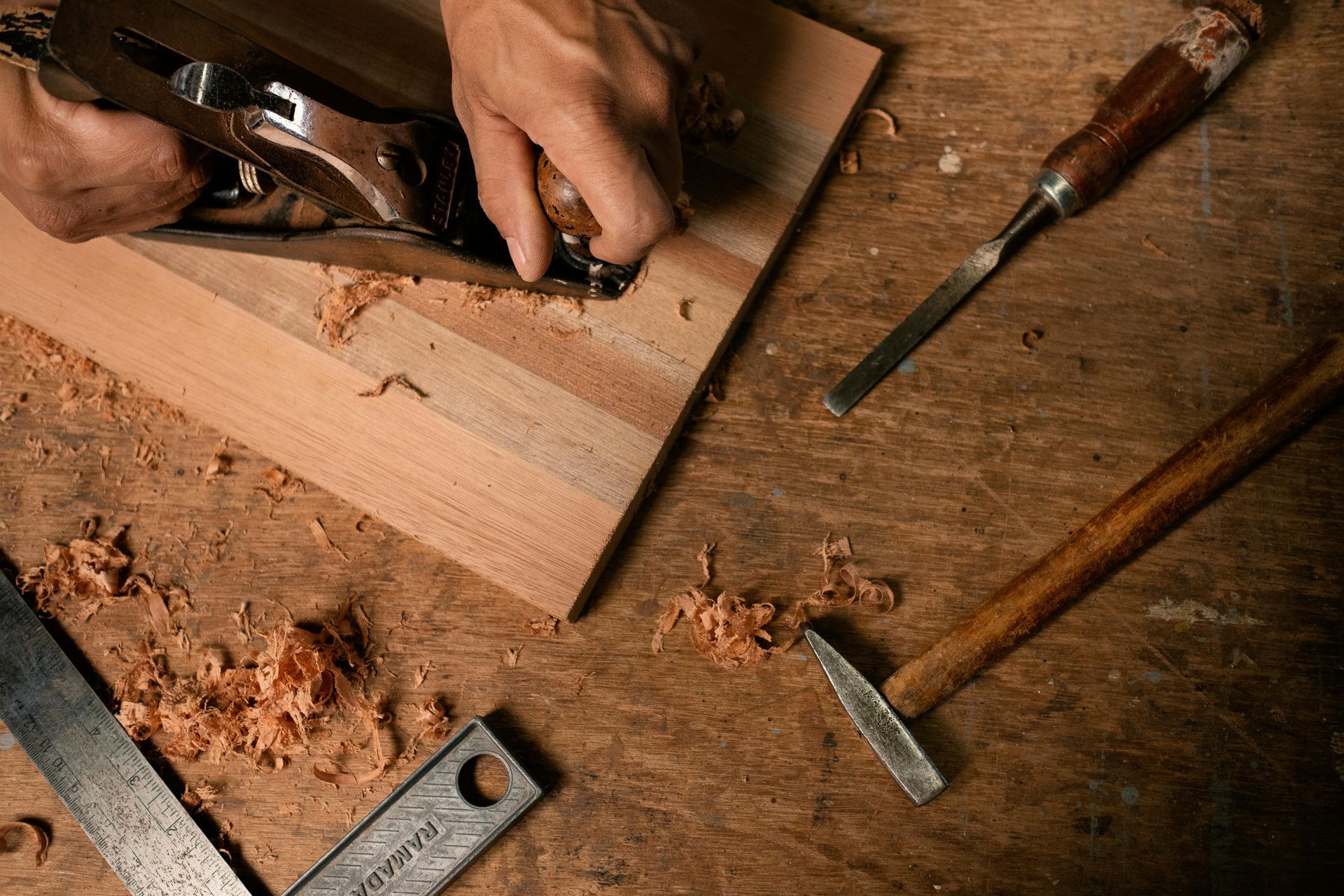 Hands using a hand plane on wood beside chisels and sawdust in a woodworking workshop