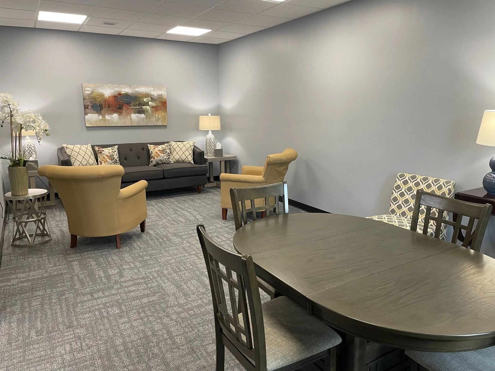 A furnished waiting room with seating, a sofa, and an oval table. Gray walls and carpet.