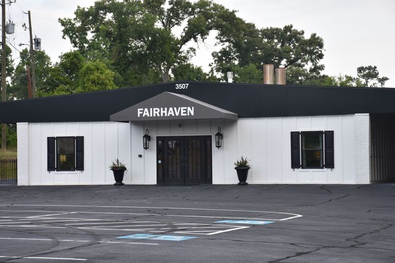 White building with black roof and double doors; two windows; asphalt parking area.