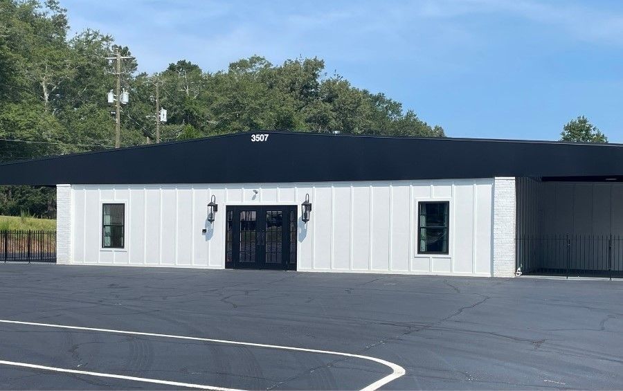 White building with black roof and double doors; two windows; asphalt parking area.