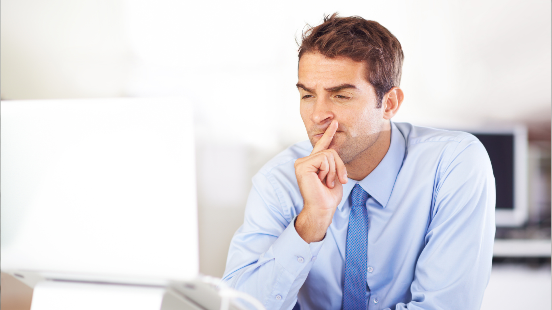 Man in blue shirt and tie at a laptop, holding finger to his nose, looking thoughtful.