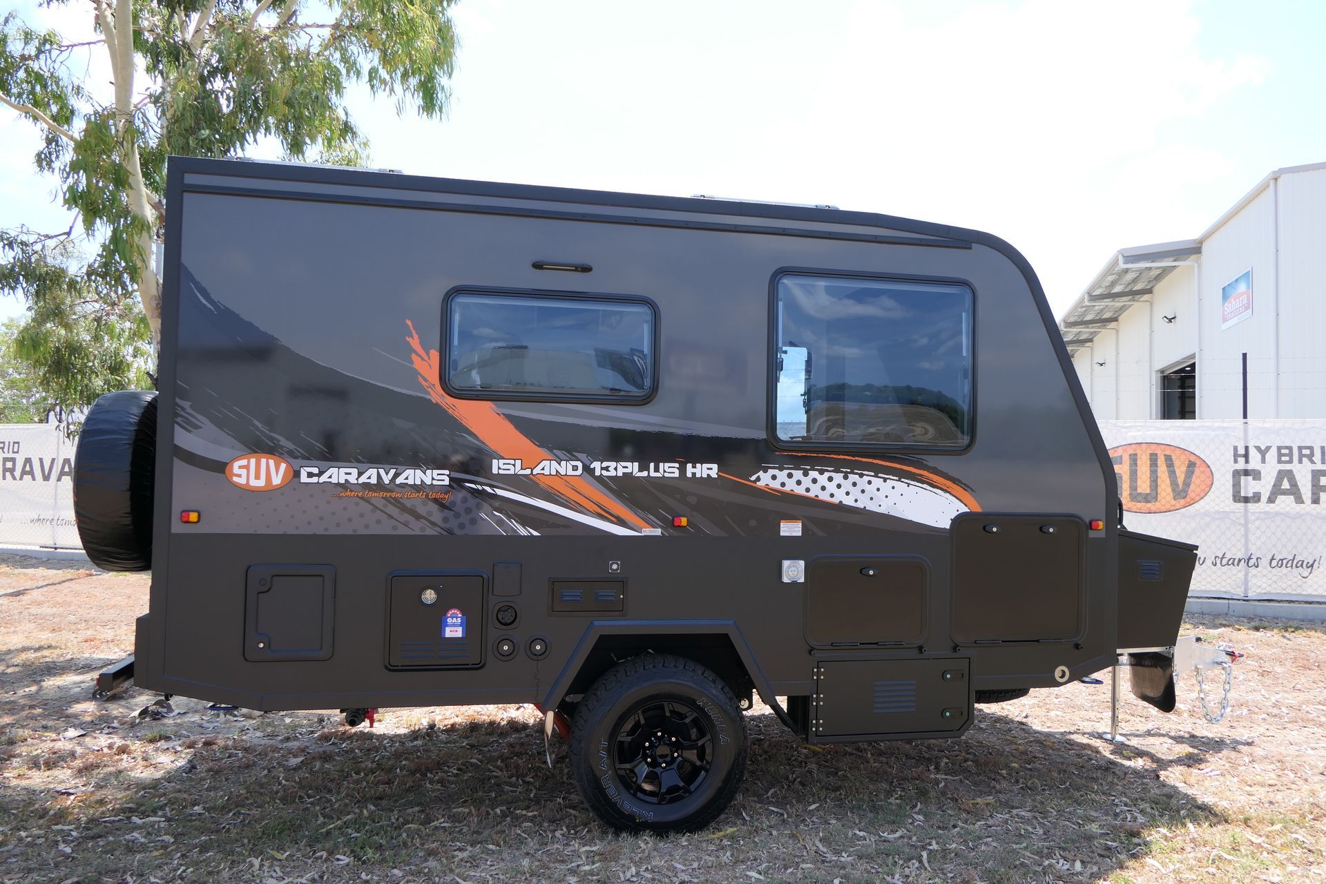 A black camper trailer is parked in a gravel lot in front of a building.