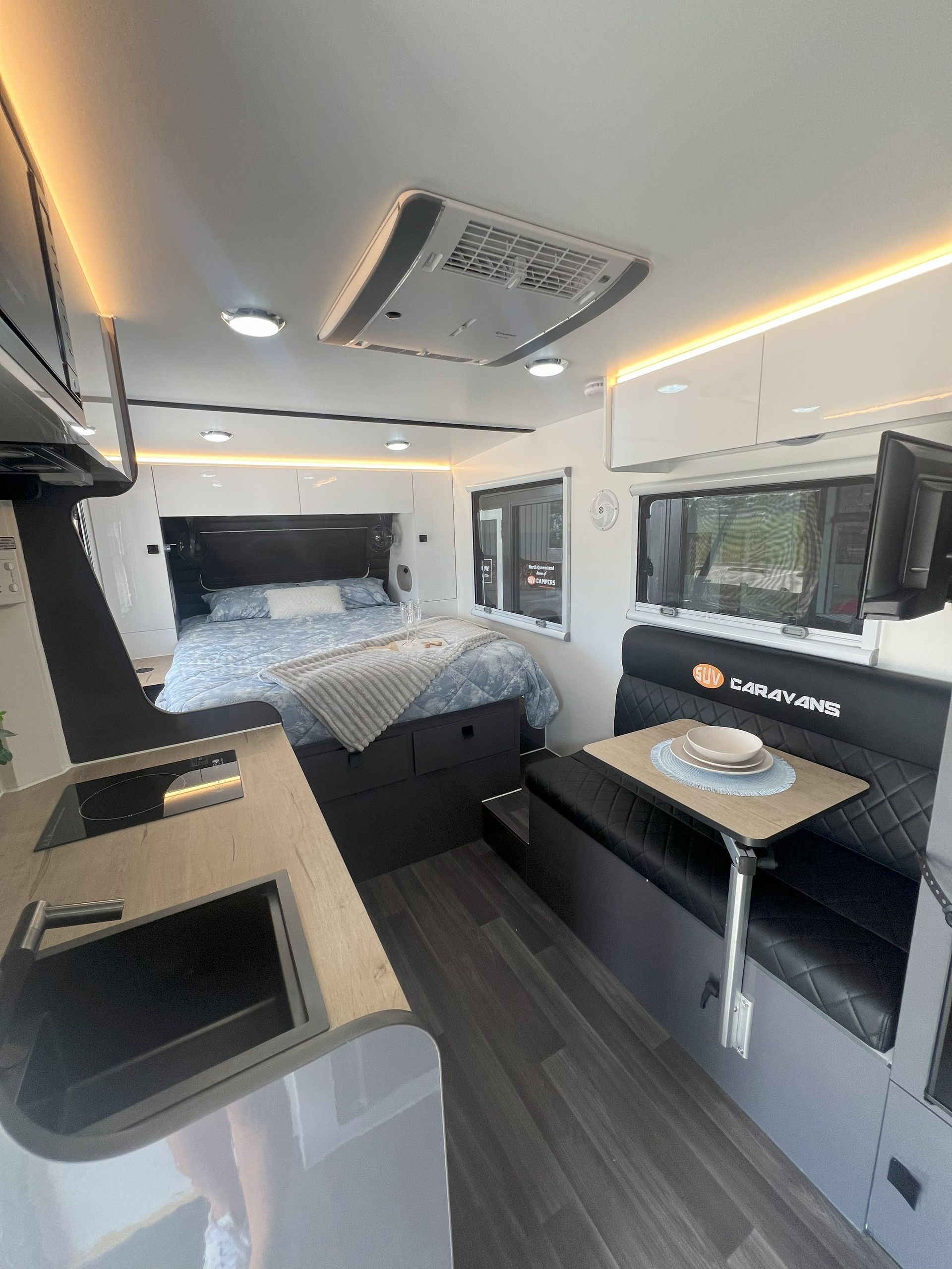 Interior of a modern caravan with a bed, seating area, kitchen, and television. Grey, white, and black color scheme.