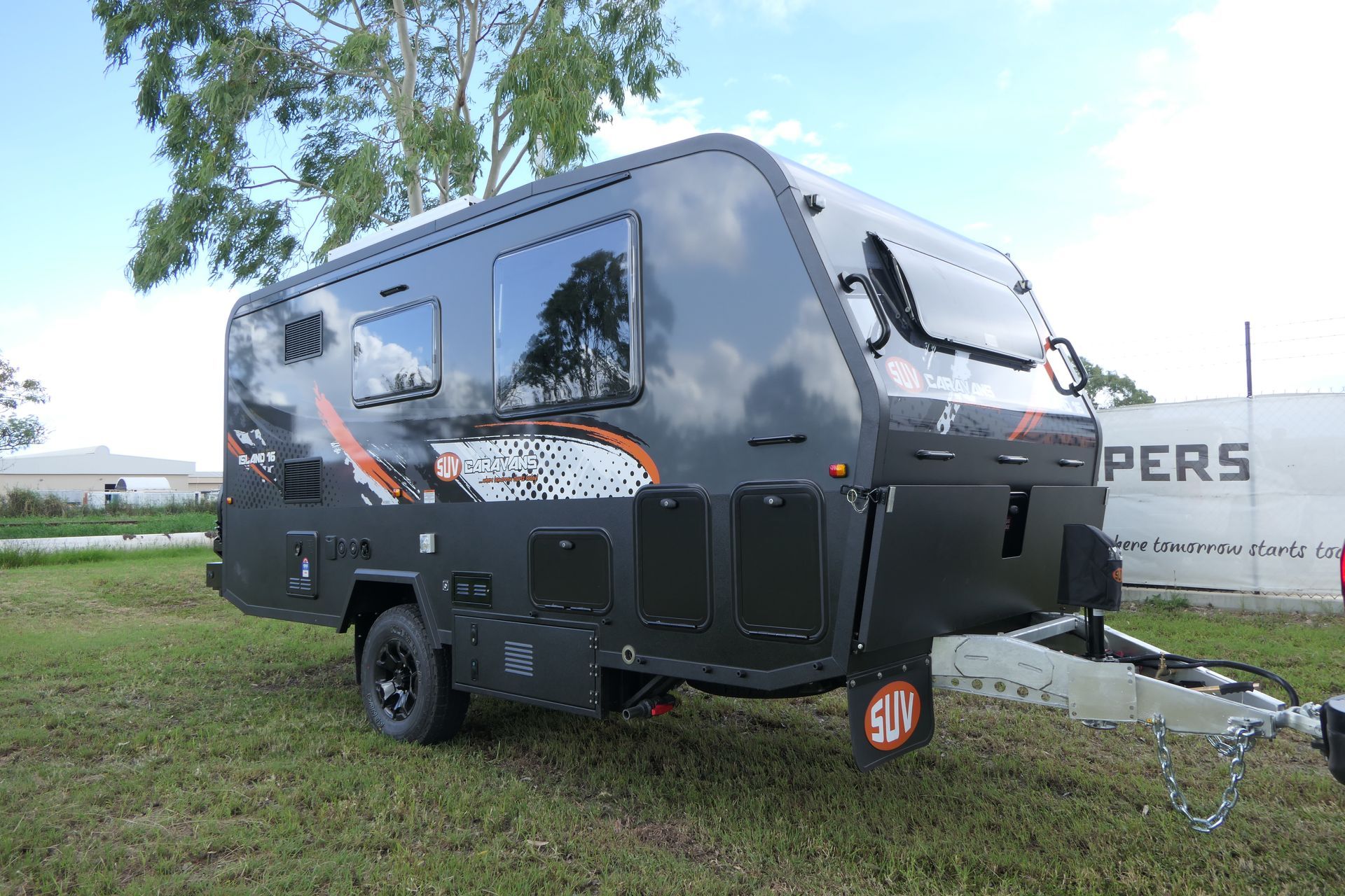 Dark gray caravan on grass with orange and white accents, under a blue sky.