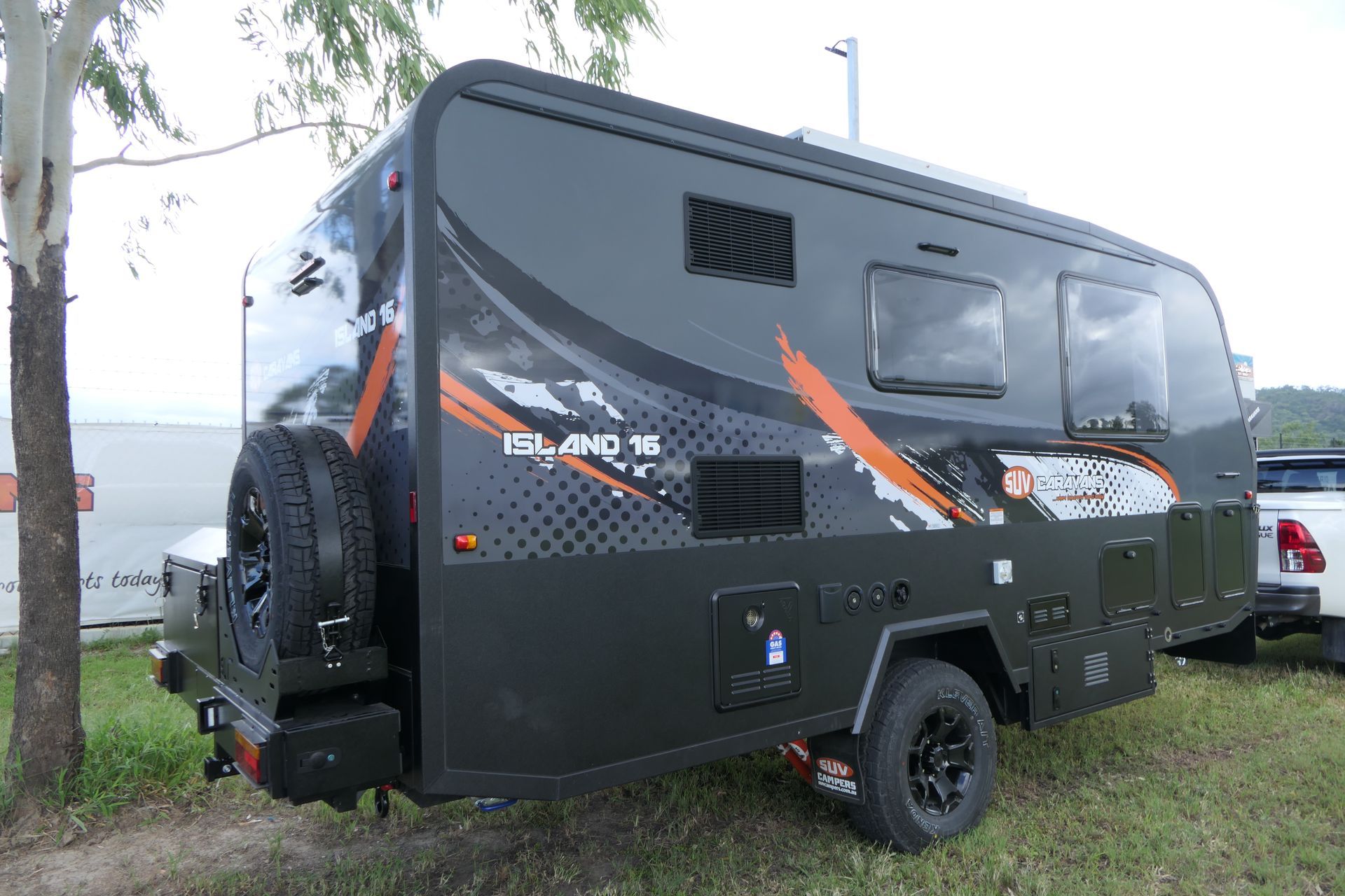 Dark gray travel trailer with orange accents parked on grass. Spare tire on back, near a tree.