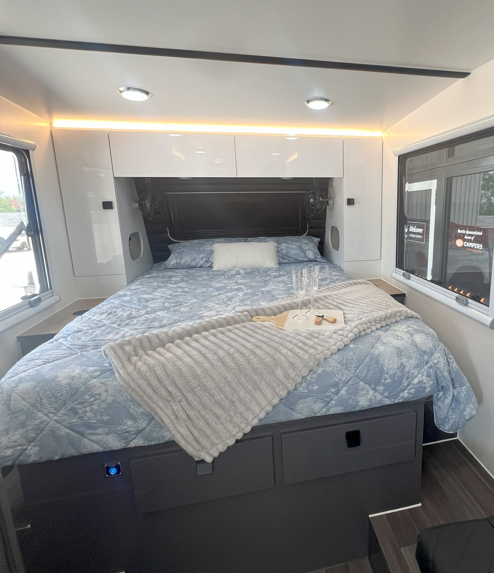 Bedroom in a caravan with a bed, comforter, and storage drawers. There are windows and overhead lights.