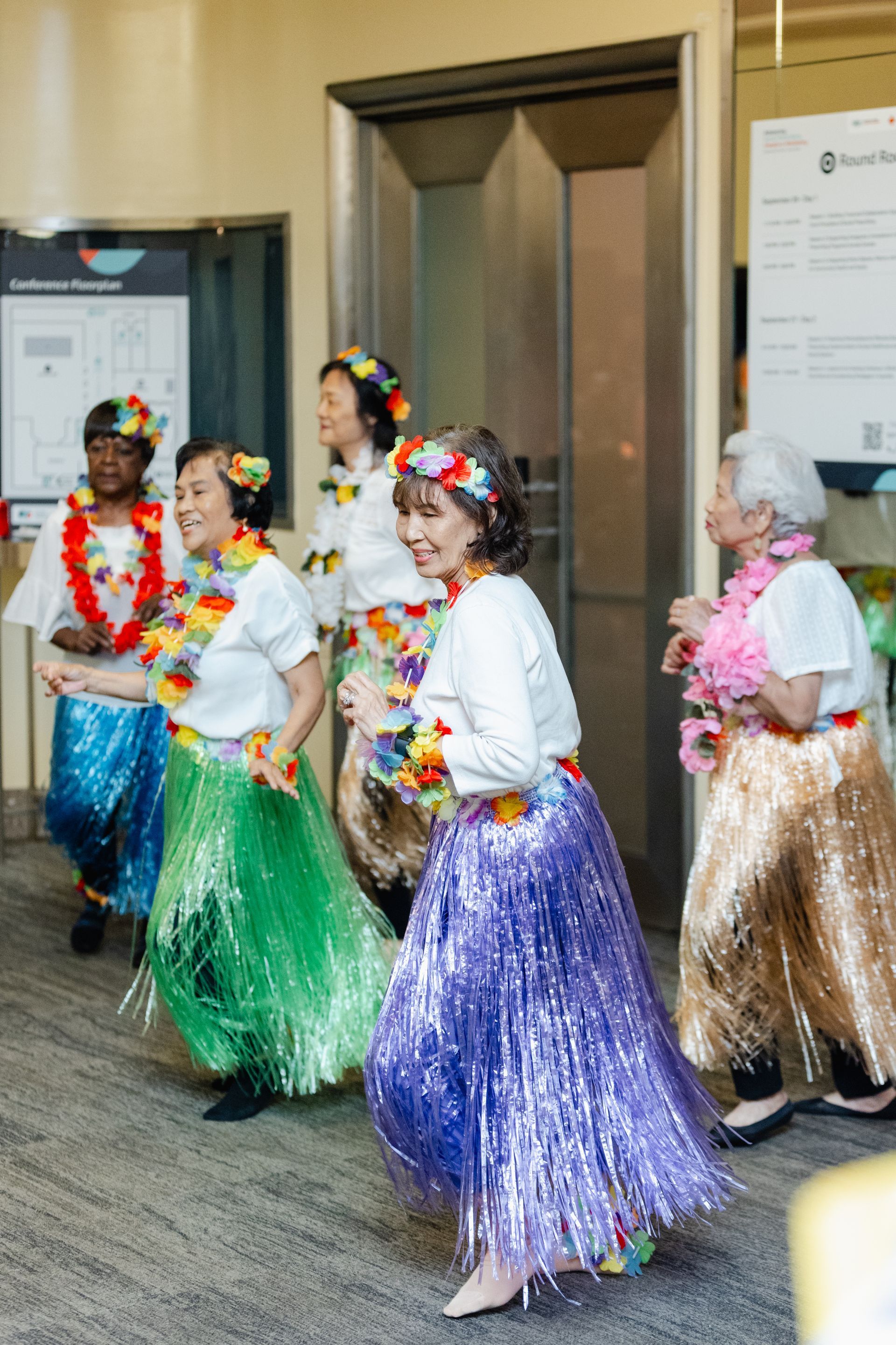 A group of elderly women are dancing hula in front of an elevator.