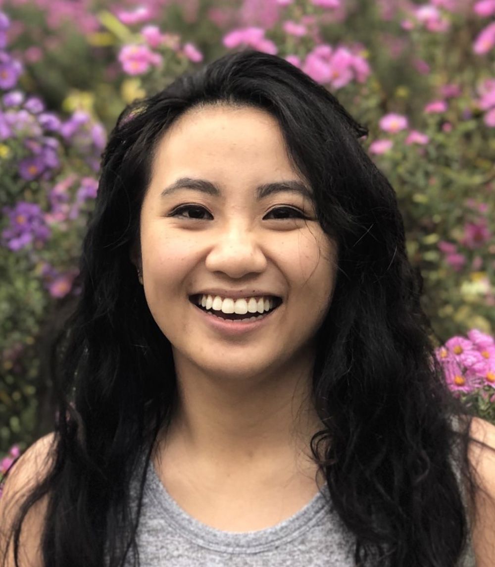 A woman is smiling in front of a bush of purple flowers.