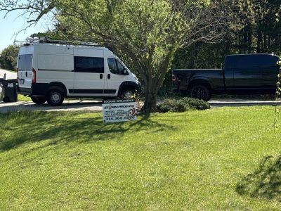 White van and black pickup parked by a tree in a grassy yard with a small sign in front.