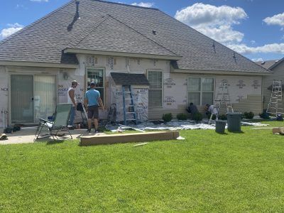 Two workers painting a house exterior with ladders and tools in a sunny backyard.