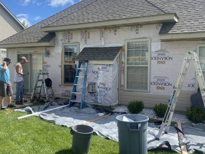 Workers set up ladders and tarps for house painting on a beige home exterior.