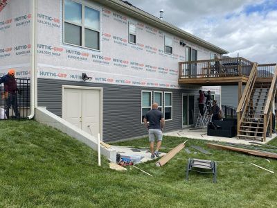Workers on a house exterior, with siding under construction, a deck, and stairs beside a grassy yard