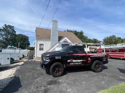 Black pickup truck with racing decals parked in a gravel lot beside a white house and trailer.