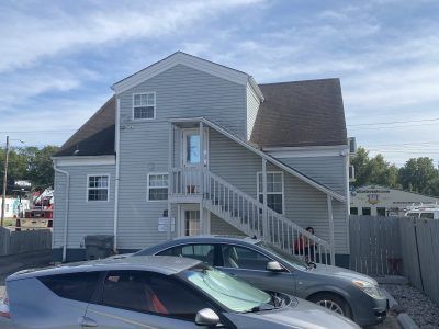 Gray two-story house with exterior staircase, parked cars, and a fenced yard under a cloudy sky