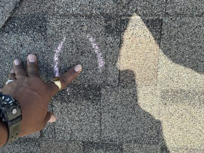Hand pointing at faint chalk markings on rough pavement beside a yellow arrow shape