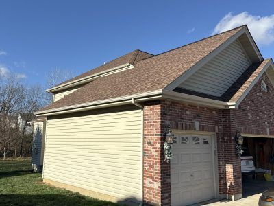 Suburban house exterior with tan siding, brick garage, and sloped shingle roof under a blue sky