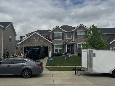 Suburban house with open garage, gray car parked in front, and a white trailer on the right under cloudy skies