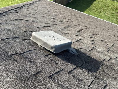 Rectangular roof vent cap on gray asphalt shingles near grassy lawn