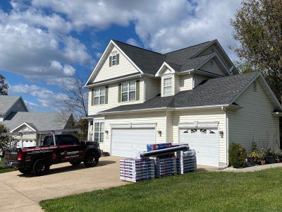 Suburban house with two white garage doors, pickup truck, and stacked building materials in the driveway.