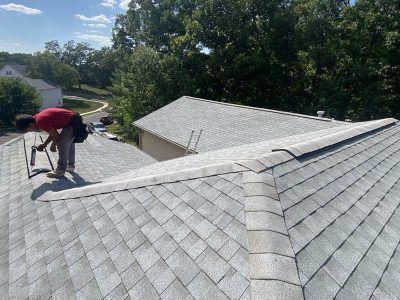 Worker on a shingled roof repairing shingles beside a tree-lined yard