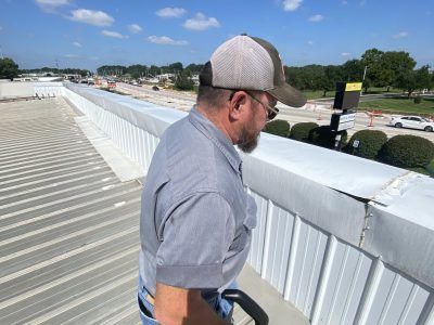 Man leaning over a white rooftop railing under a blue sky, looking down at the ground below