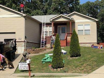 Workers repairing a beige house roof and porch, with ladders, tools, and a dumpster in the yard