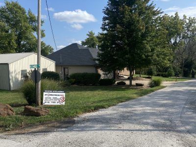 Gravel driveway beside a house and garage, with trees, lawn, and a small roadside sign.