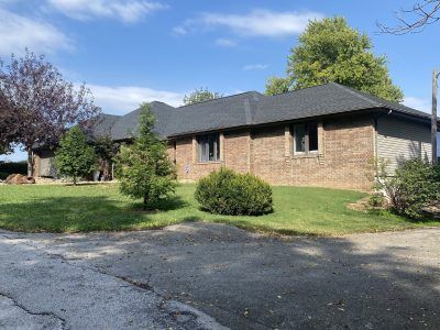 Single-story brick house with dark roof, front lawn, and cracked driveway under a blue sky.