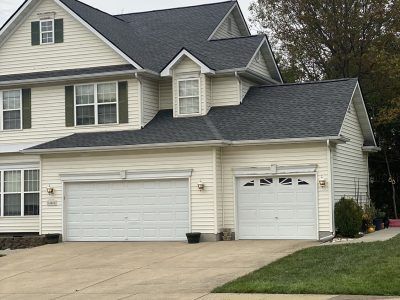 Suburban two-story house with beige siding, dark roof, and three-car garage viewed from the driveway