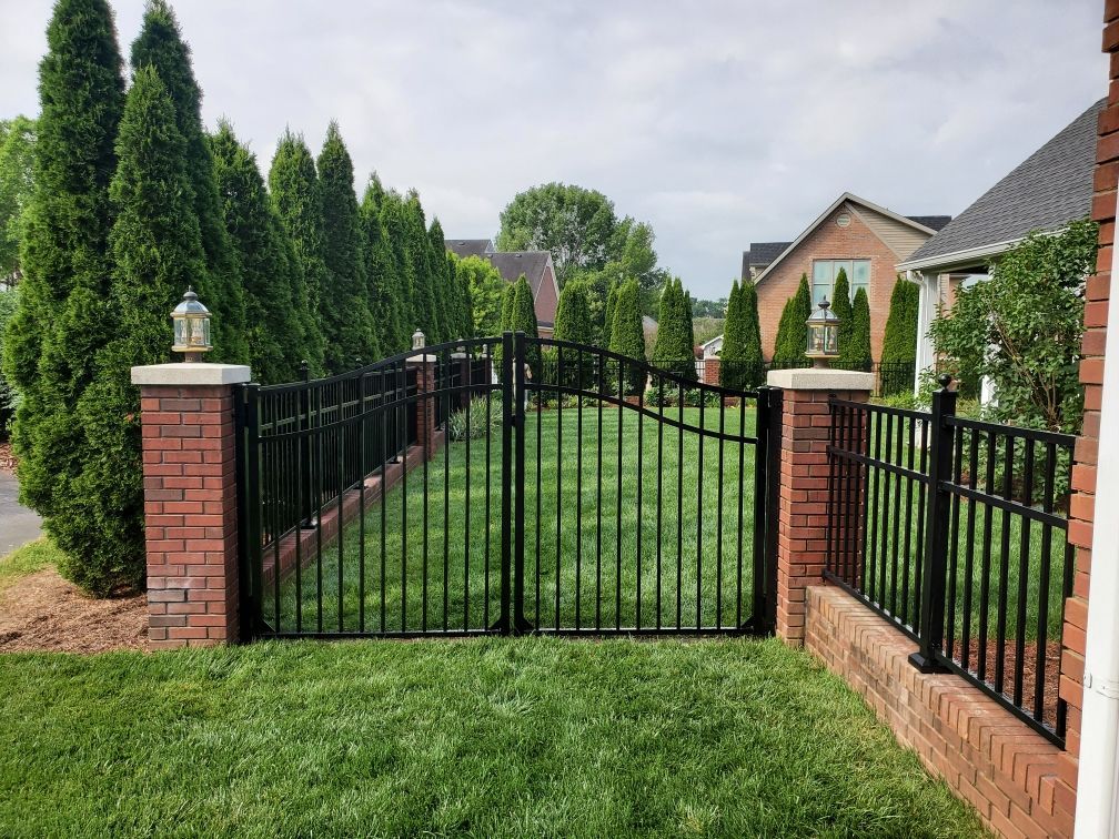 Black wrought iron gate and fence with brick pillars and lamps, bordering a grassy lawn.