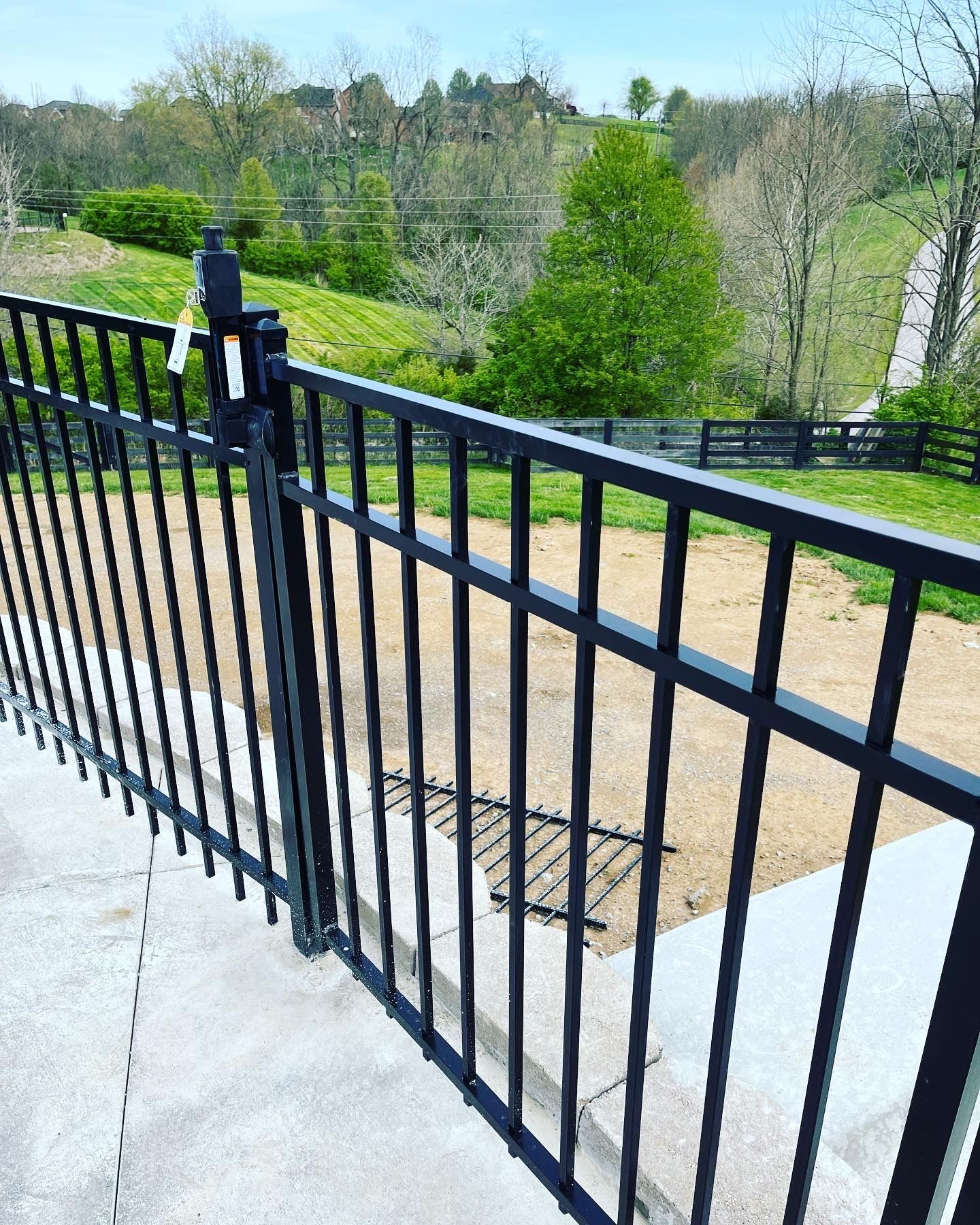Black metal fence with gate overlooking a green hillside with trees and a pathway.