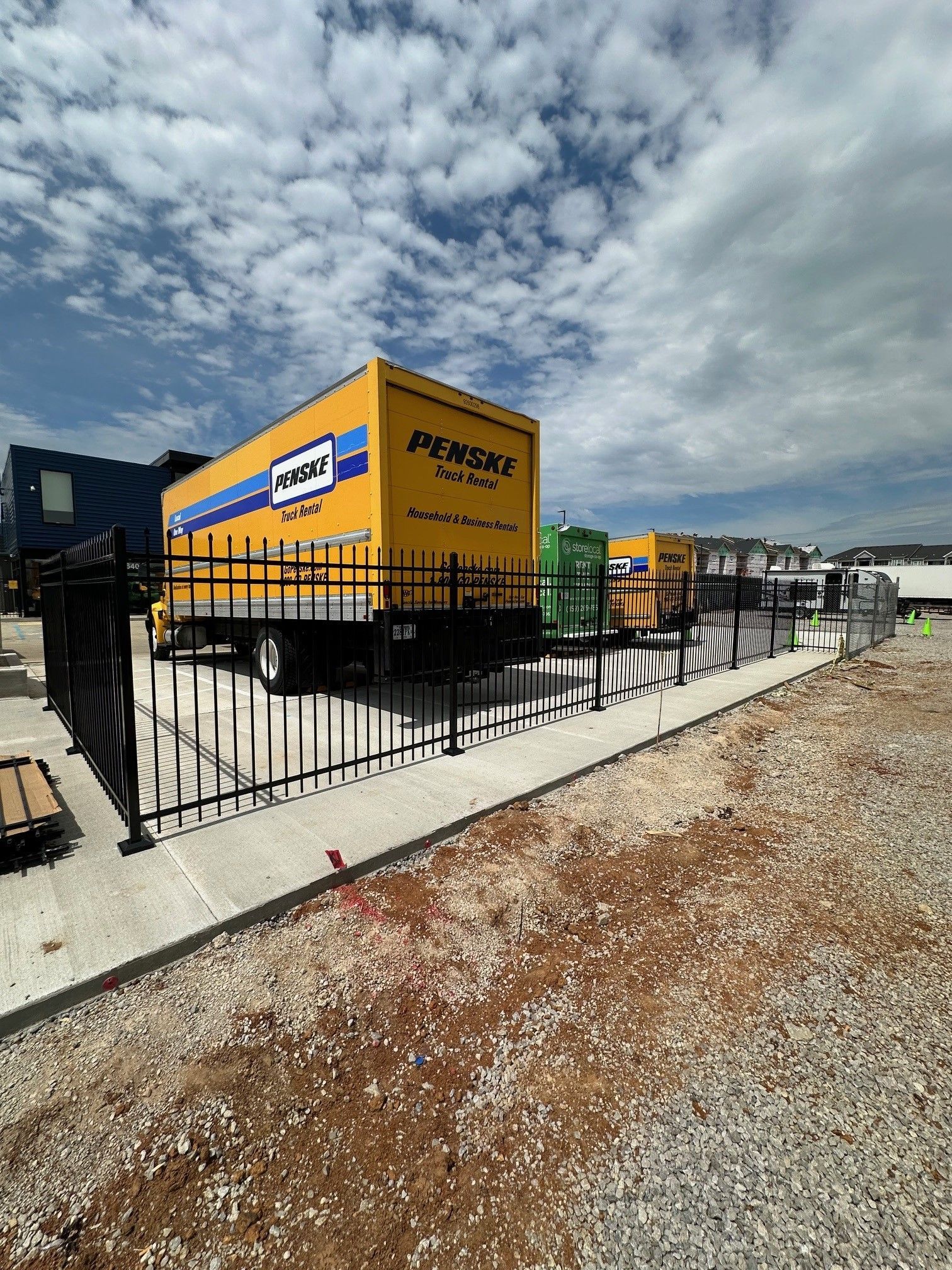 A row of moving trucks are parked in a gravel lot behind a metal fence.