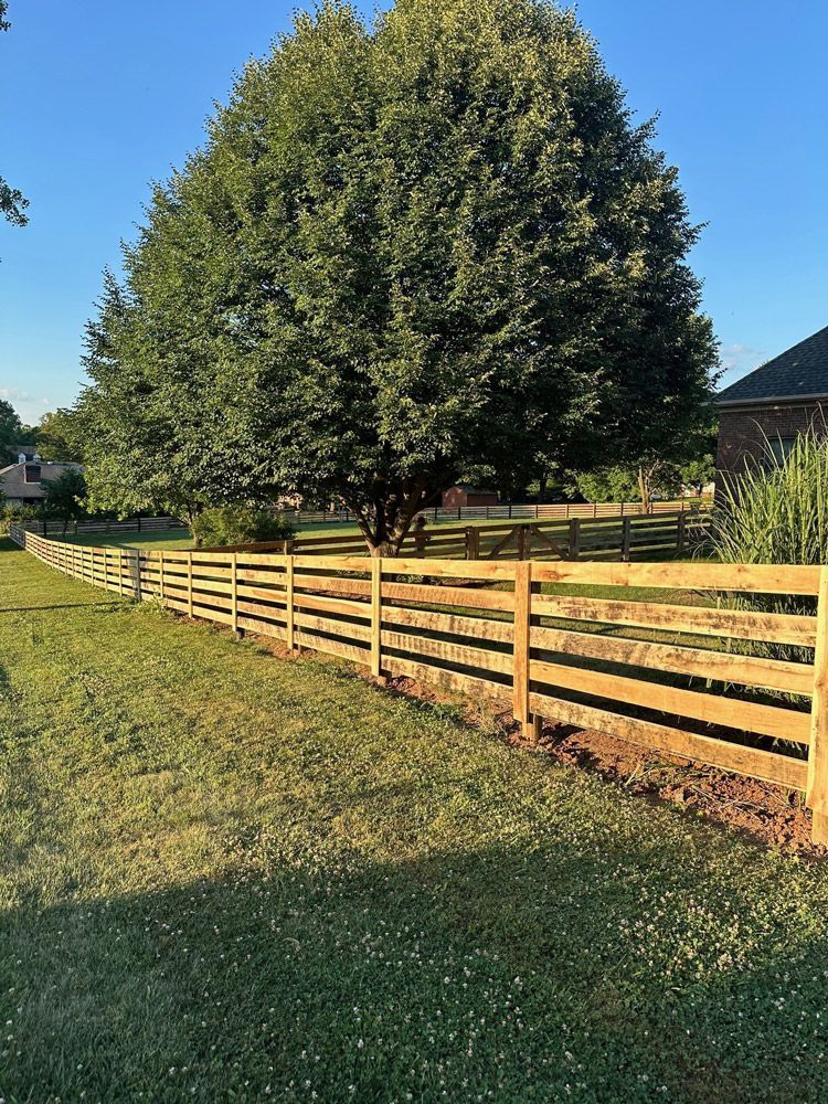 A wooden fence surrounds a lush green field with a large tree in the background.