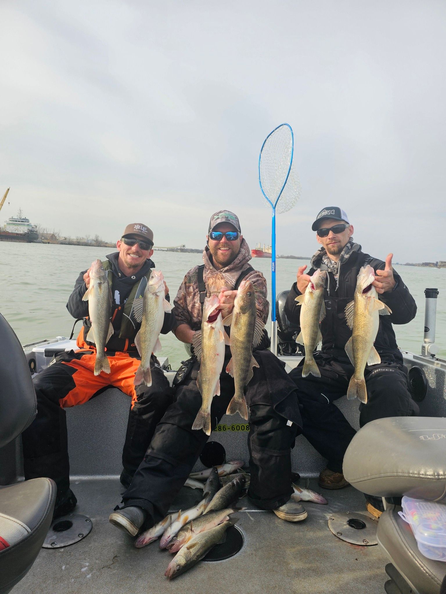Three anglers on a boat hold up fresh-caught fish, with more fish on the deck and water behind them.