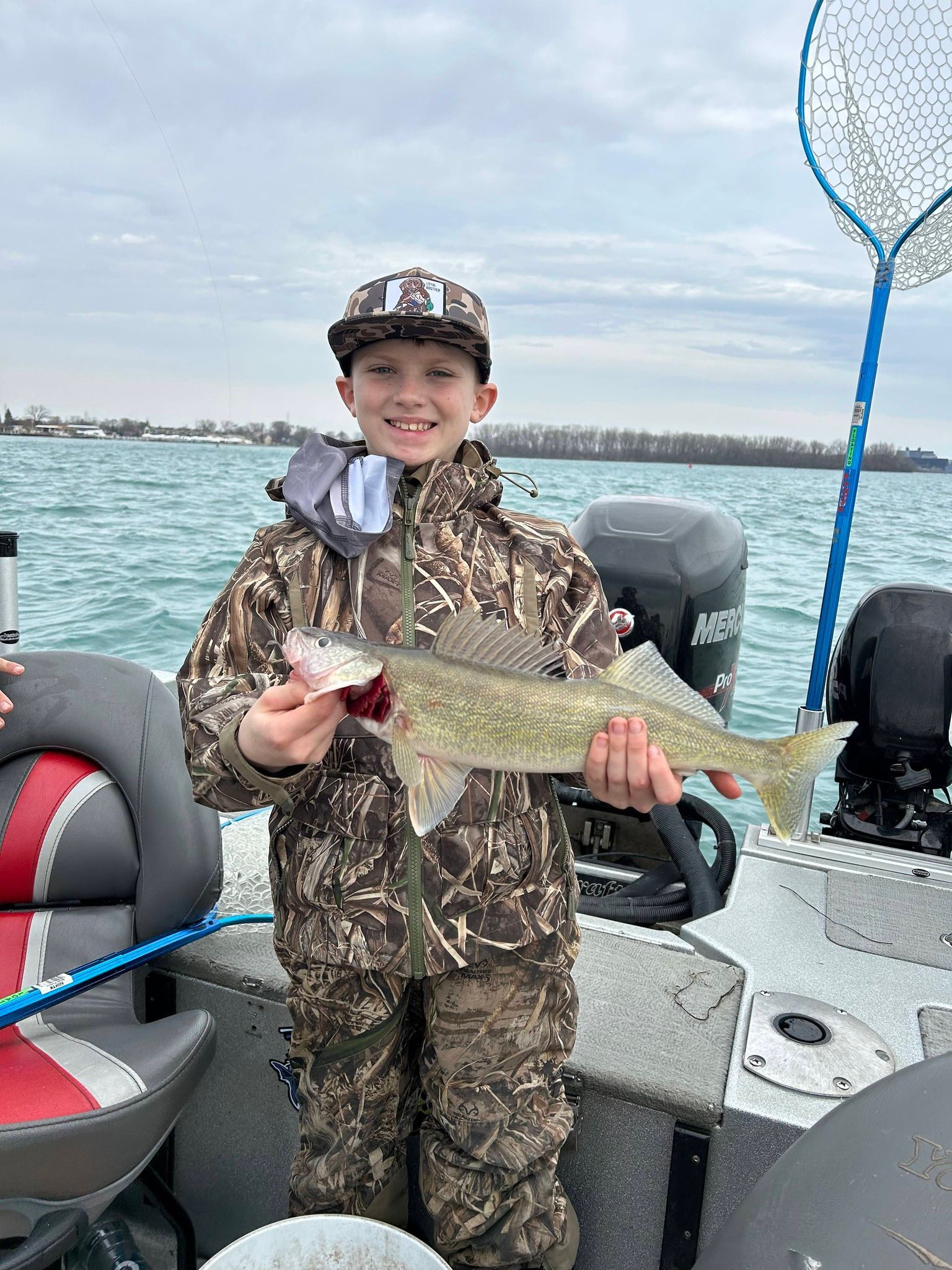 Person on a boat holding a fish, wearing camouflage, with water and shoreline in the background