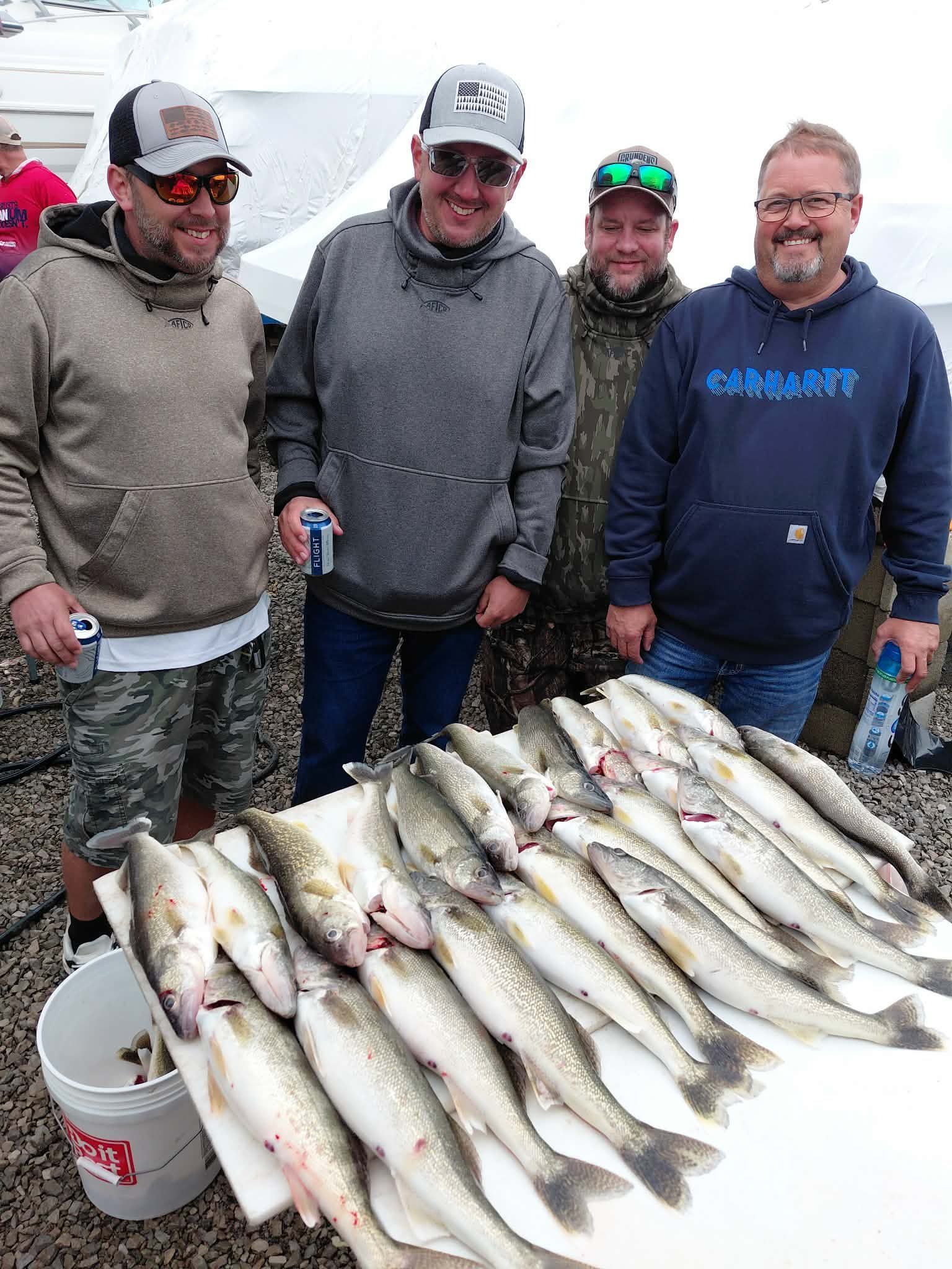 Four people stand behind a table covered with several freshly caught fish and a bucket outdoors.