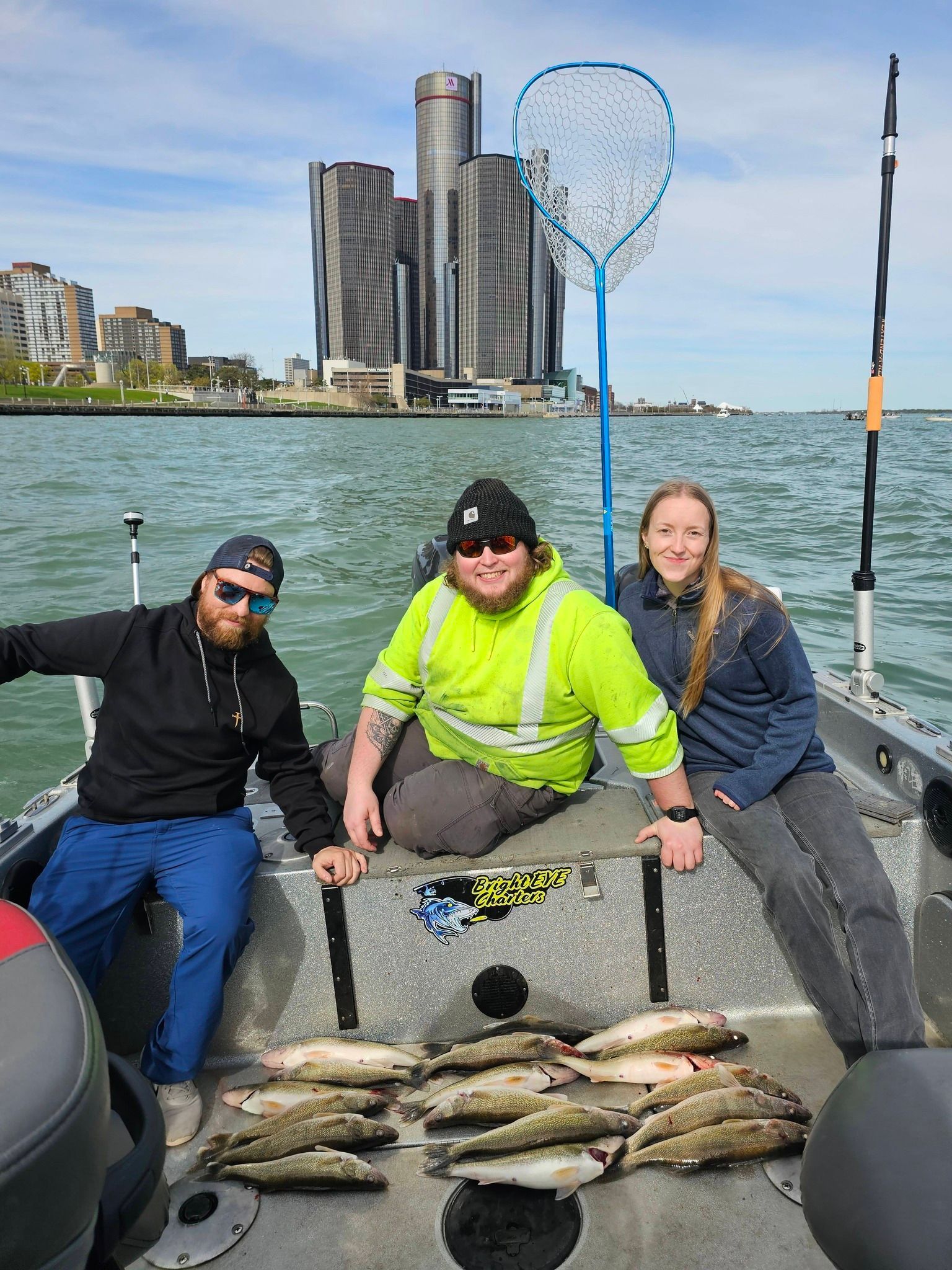 Three people on a fishing boat with several fish laid out, city skyline and water in the background.