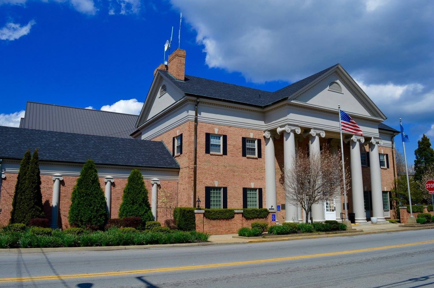 Brick civic building with white columns, American flags, and landscaped entrance under a blue sky