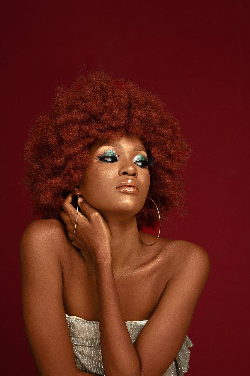 A woman with red hair and blue eyeshadow is standing in front of a red background.