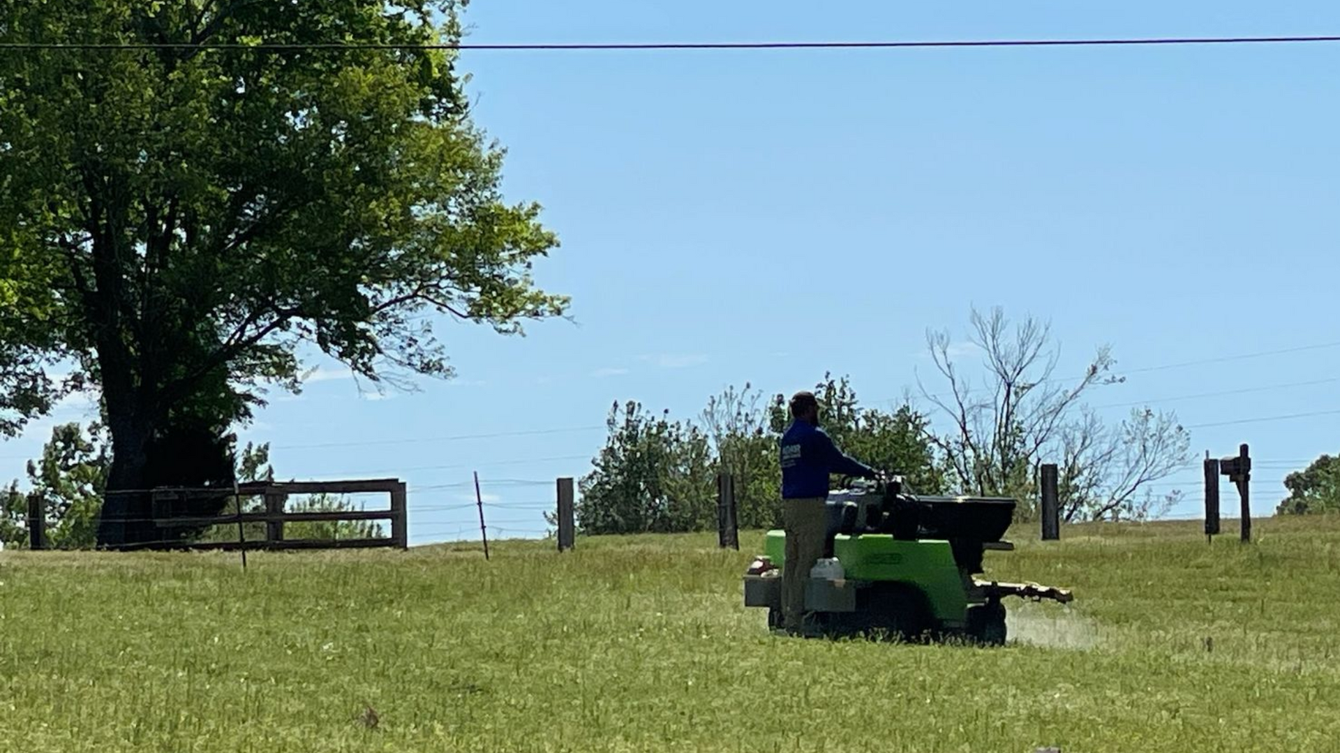 A person rides a green utility vehicle across a grassy field near a large tree and a wooden fence on a sunny day.