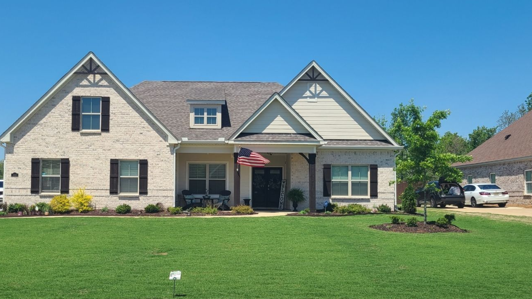 A two-story house with cream brick, a dark shingled roof, a front porch with a flag, and a grassy front yard.