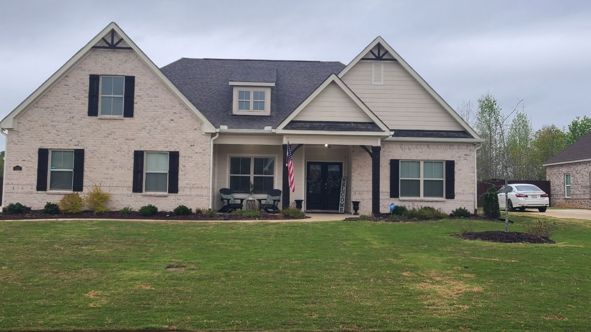 A light-colored brick, two-story house with black shutters, a covered porch, and a lawn under a cloudy sky.