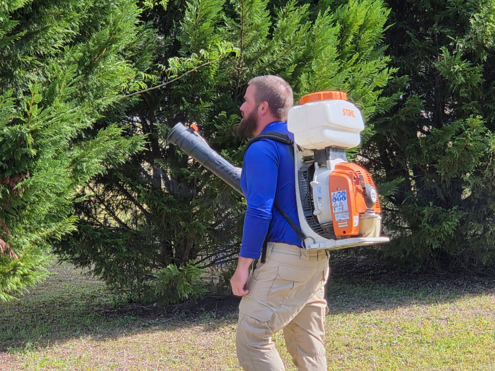 A person in a blue long-sleeved shirt and tan pants wears a backpack blower, directing its nozzle toward an evergreen tree.